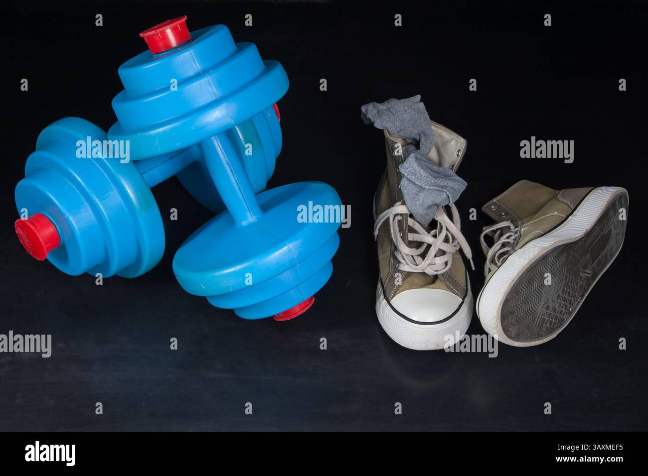 On the scratched rubber floor of an empty gym lie blue dumbbells and worn-out sneakers with old socks. Stock Photo