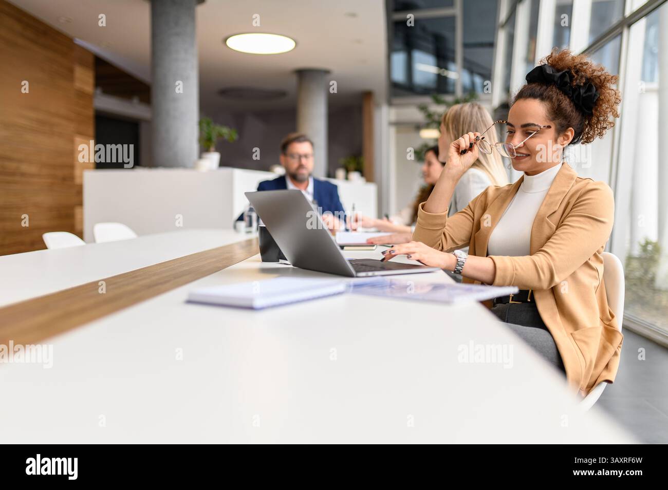 Businesswoman wearing eyeglasses while analyzing data over laptop in office with coworkers in the background Stock Photo