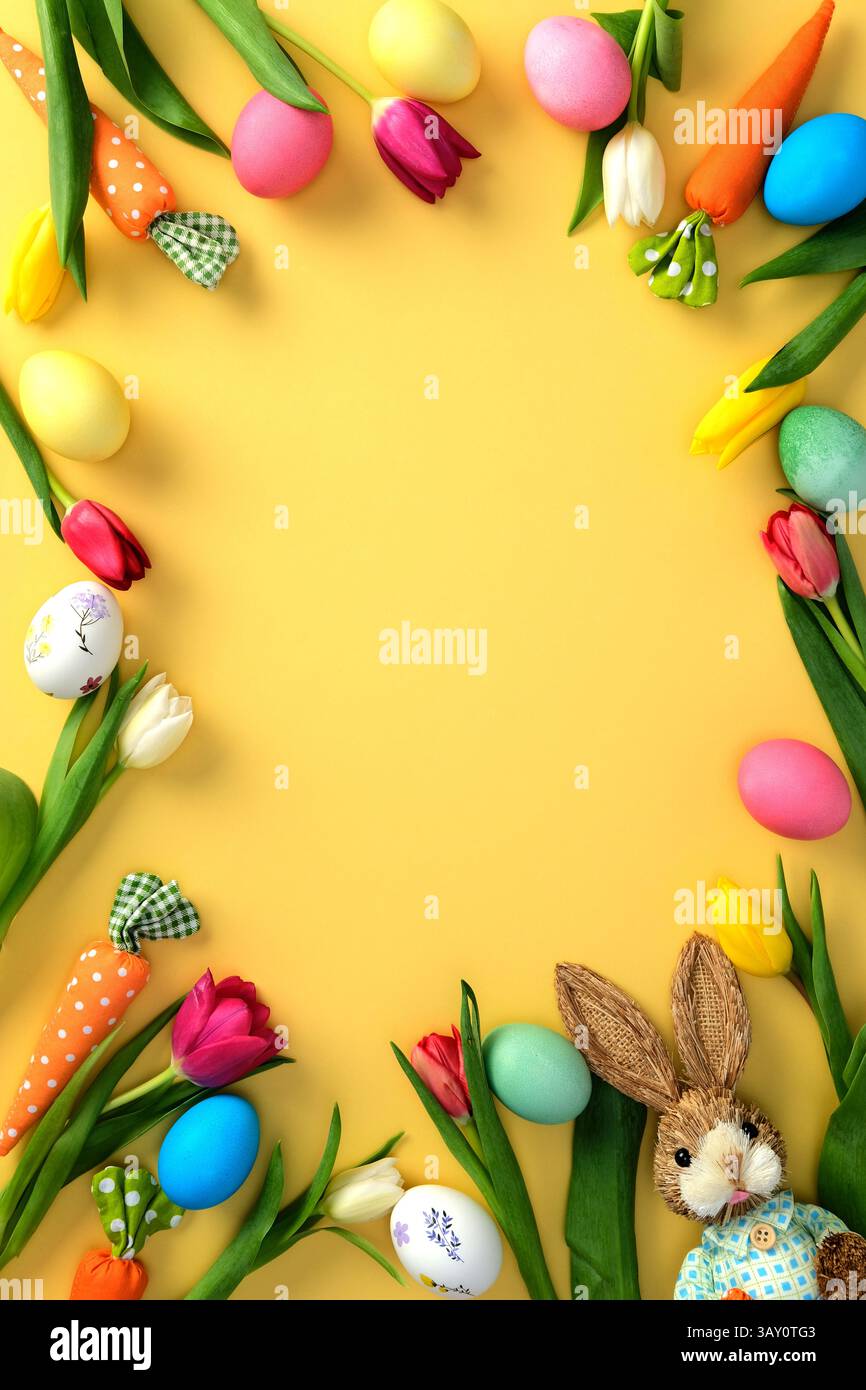 Frame made of painted Easter eggs, festive decorations, and tulip flowers on yellow background. Stock Photo