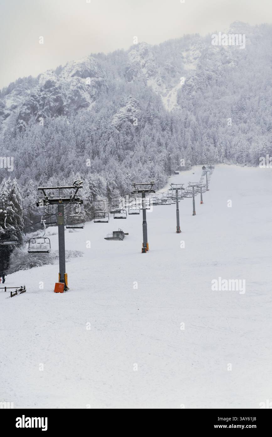 A snowy ski slope stretches through a wintery forest, with skiers descending amidst snow-covered trees and lift infrastructure Stock Photo
