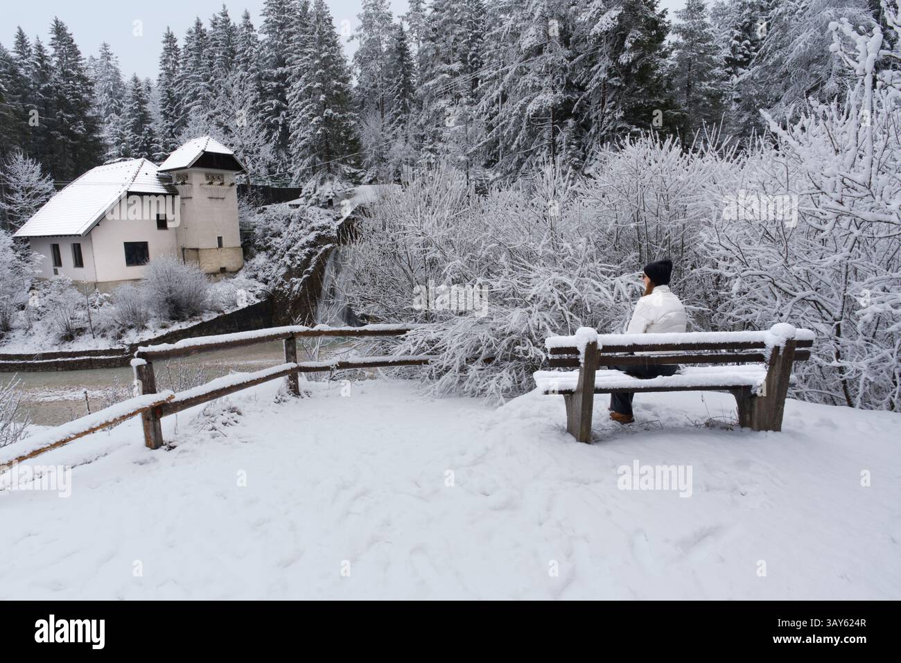A girl sits peacefully on a snow-covered bench, surrounded by a serene winter landscape of snowy trees, a distant house, and a wooden fence. Slovenia Stock Photo