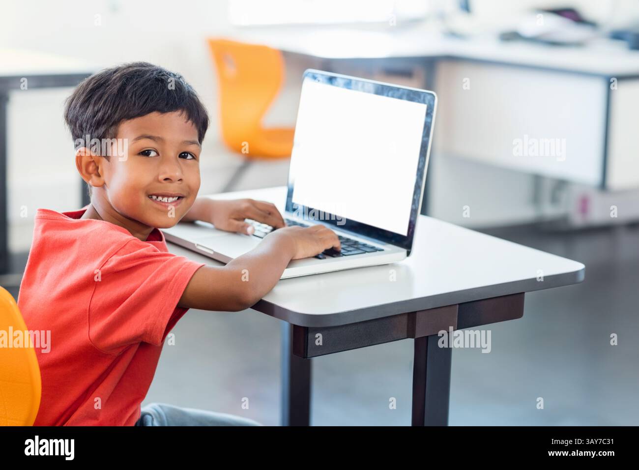 Boy typing on laptop at desk smiling at camera in classroom, with orange chairs, copy space Stock Photo