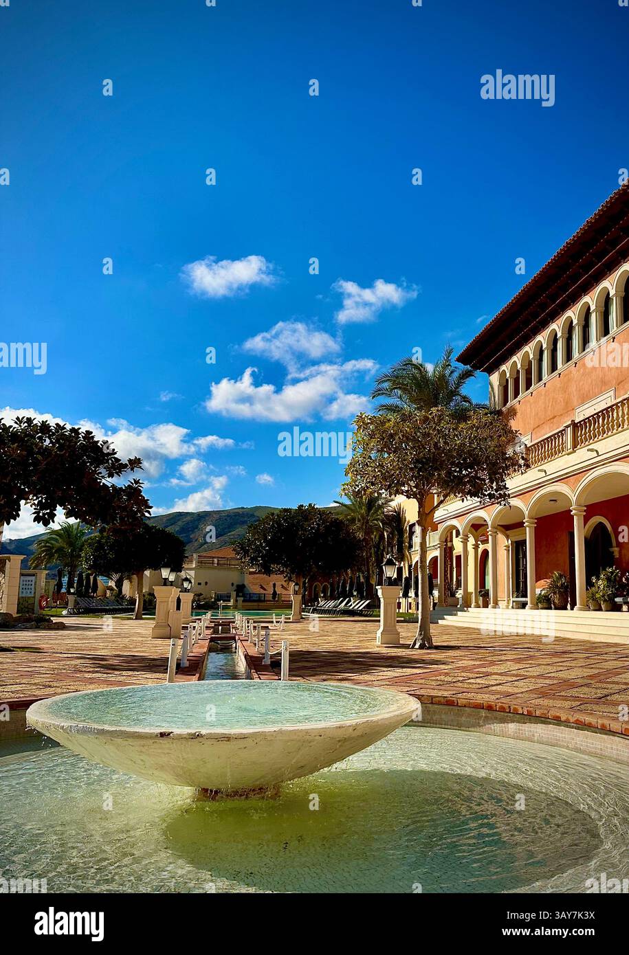 This elegant scene captures a sunny courtyard of a luxury resort, with a serene fountain as the focal point. The flowing water adds a sense of tranqui - Smartphone Captured Stock Image