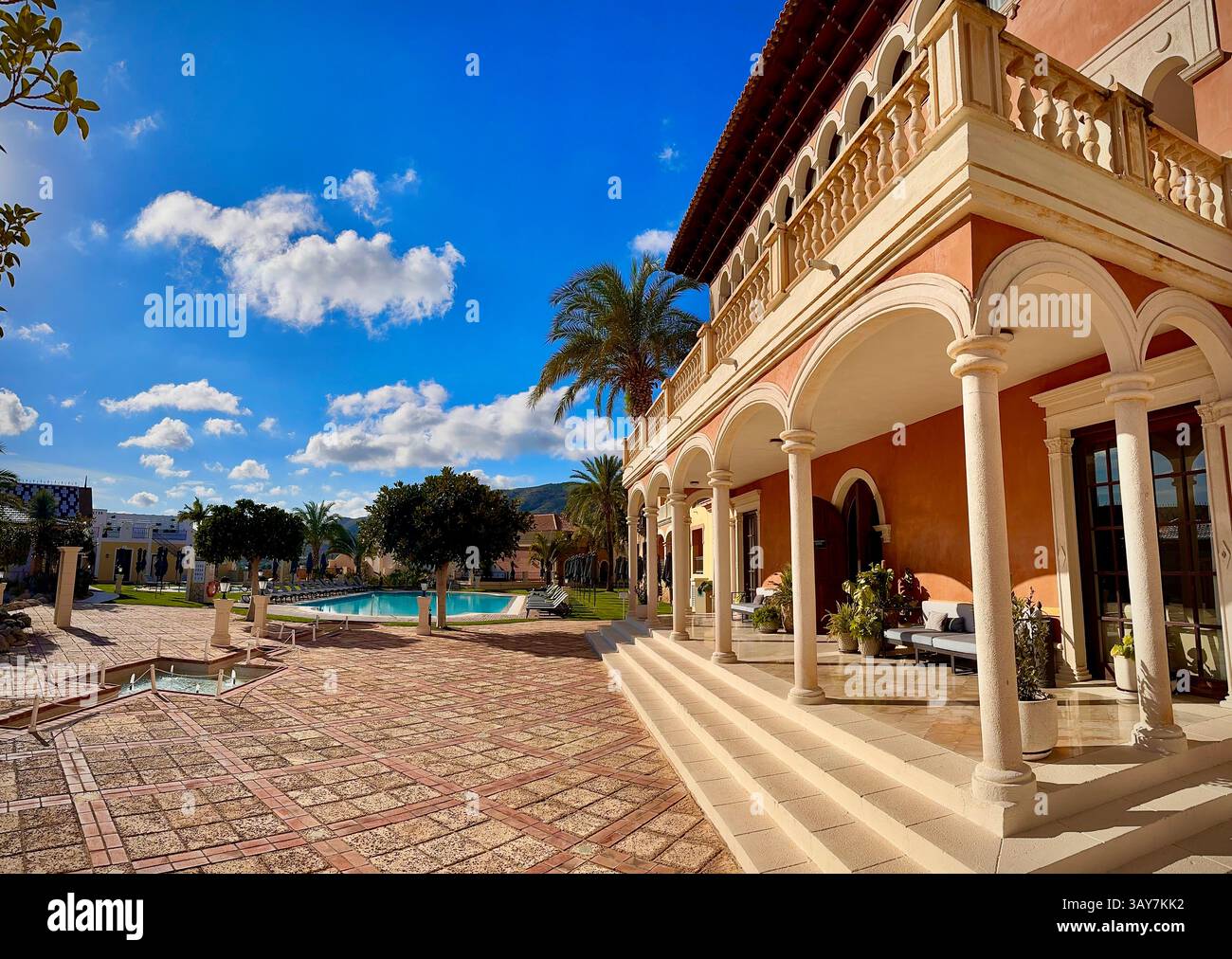 This picturesque scene showcases a luxury resort courtyard on a sunny day. The clear blue sky, fluffy clouds, and palm trees set the perfect backdrop - Smartphone Captured Stock Image