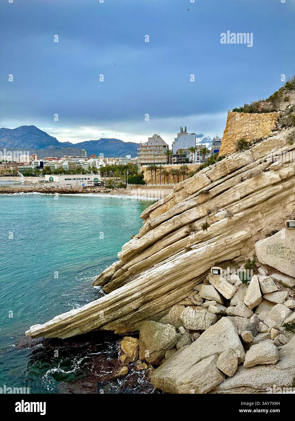 Sharp, layered rock formations meet turquoise Mediterranean waters as the city quietly rests in the background — a striking harmony of nature's raw ed - Smartphone Captured Stock Image Sharp, layered rock formations meet turquoise Mediterranean waters as the city quietly rests in the background — a striking harmony of nature's raw ed - Smartphone Captured Stock Image