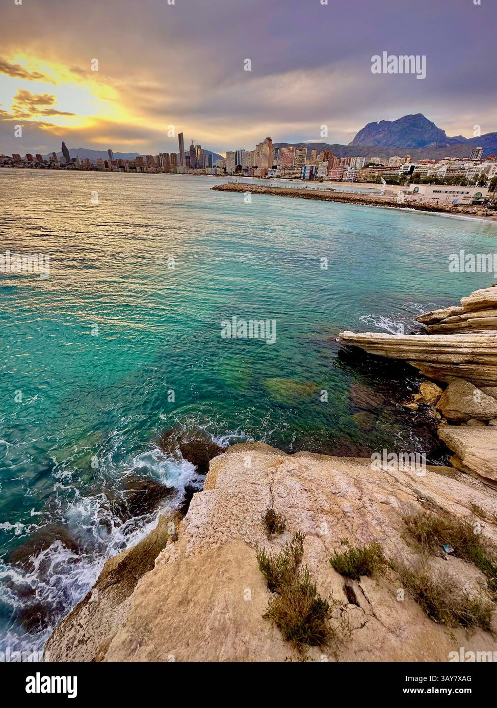 A golden sunset embraces the skyline of a Mediterranean city as the waves gently crash against rugged coastal rocks — a moment of serenity where natur - Smartphone Captured Stock Image A golden sunset embraces the skyline of a Mediterranean city as the waves gently crash against rugged coastal rocks — a moment of serenity where natur - Smartphone Captured Stock Image