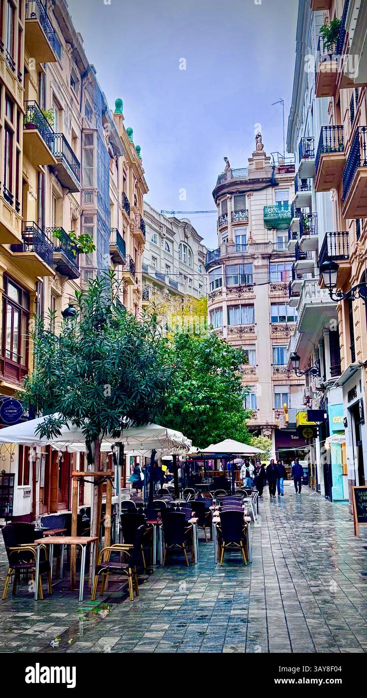 Colorful narrow street in Valencia with historic buildings, wet pavement, and outdoor cafés under umbrellas after rain. - Smartphone Captured Stock Image