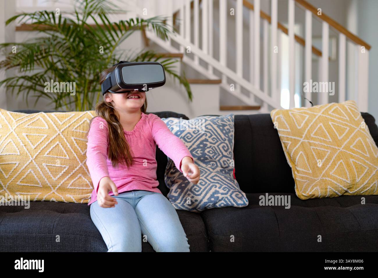 Girl sitting on dark grey couch wearing VR headset in modern living room with decorative pillows Stock Photo
