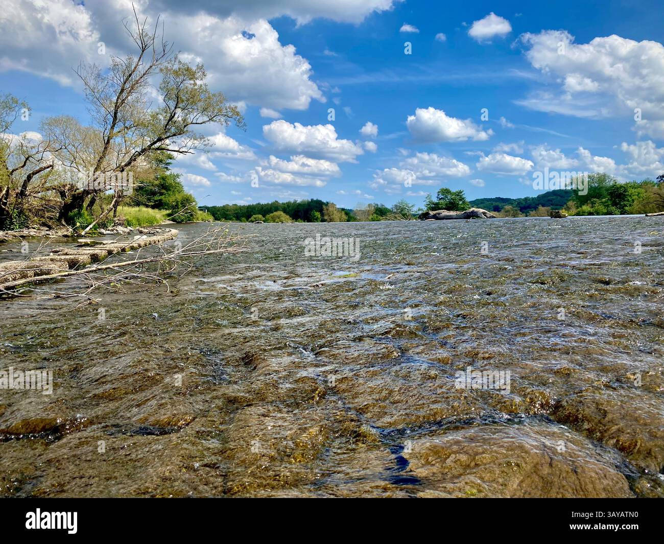 This photo captures a shallow river with crystal-clear water flowing over natural stone textures, framed by trees and greenery under a vibrant blue sk - Smartphone Captured Stock Image