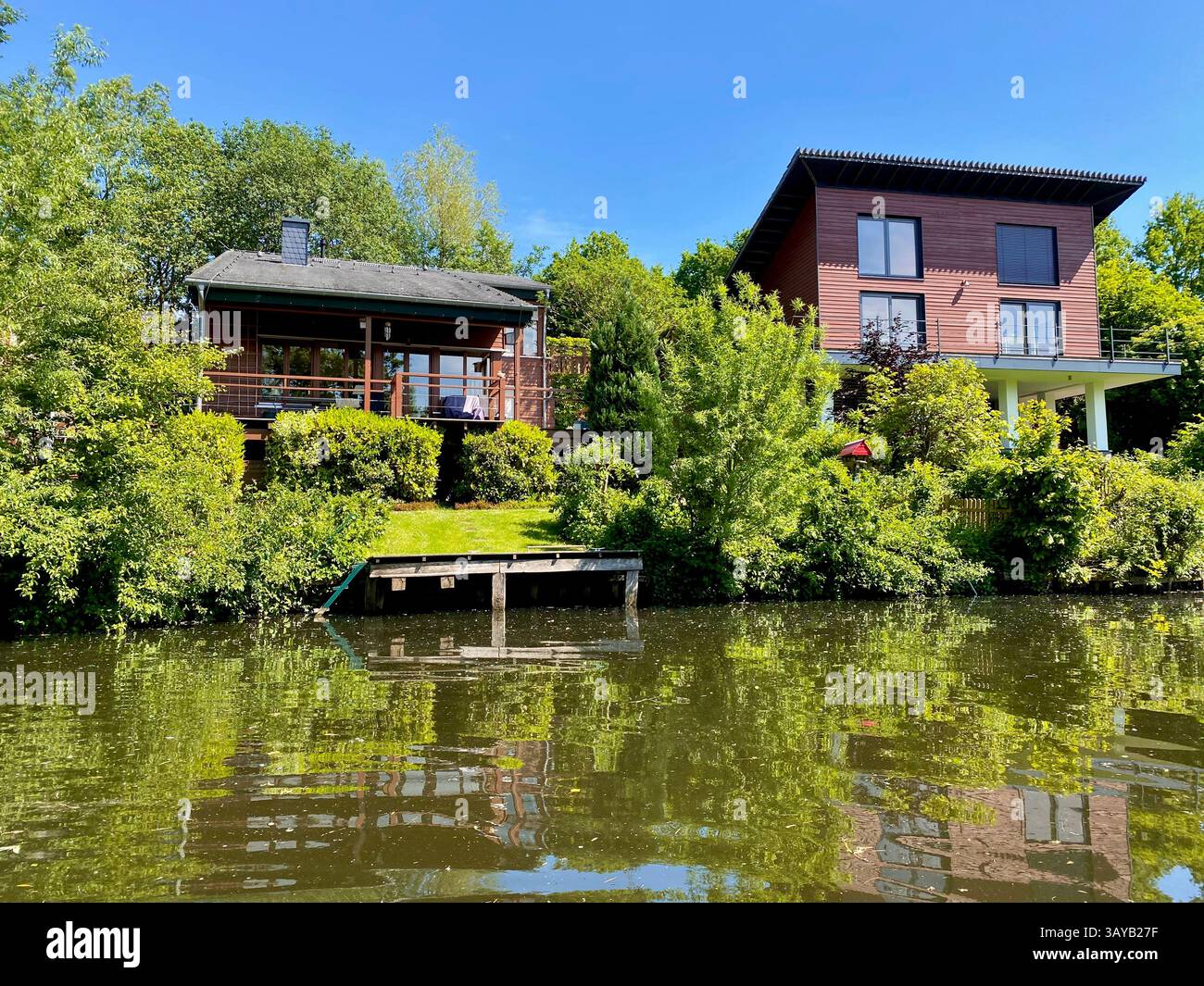 A scenic riverside view showcasing two distinct houses—one with rustic charm and the other with modern architecture—nestled in lush greenery. The calm - Smartphone Captured Stock Image