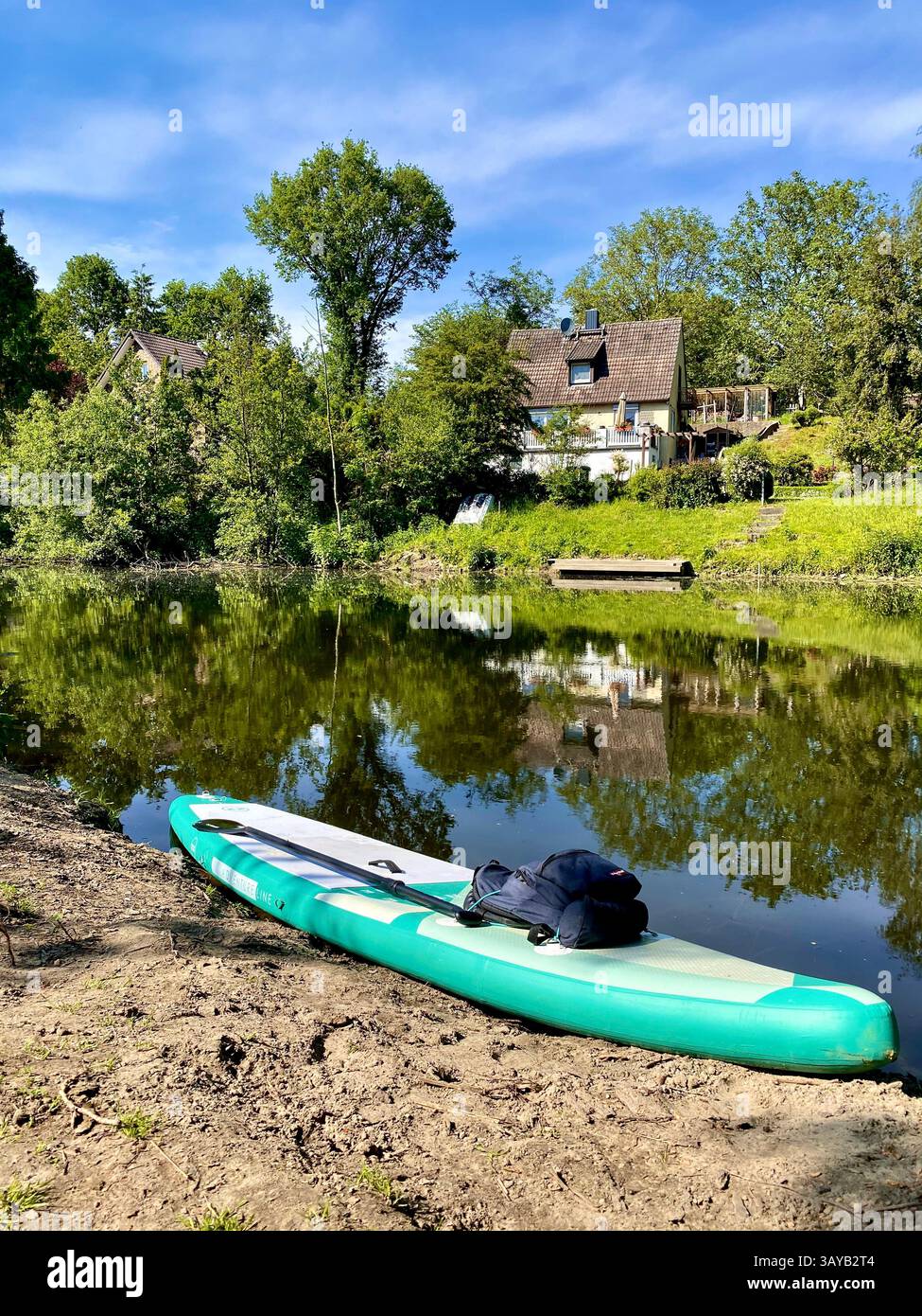A green inflatable water board lies on the sandy riverbank next to calm water. In the background, houses and trees are reflected in the surface. The s - Smartphone Captured Stock Image