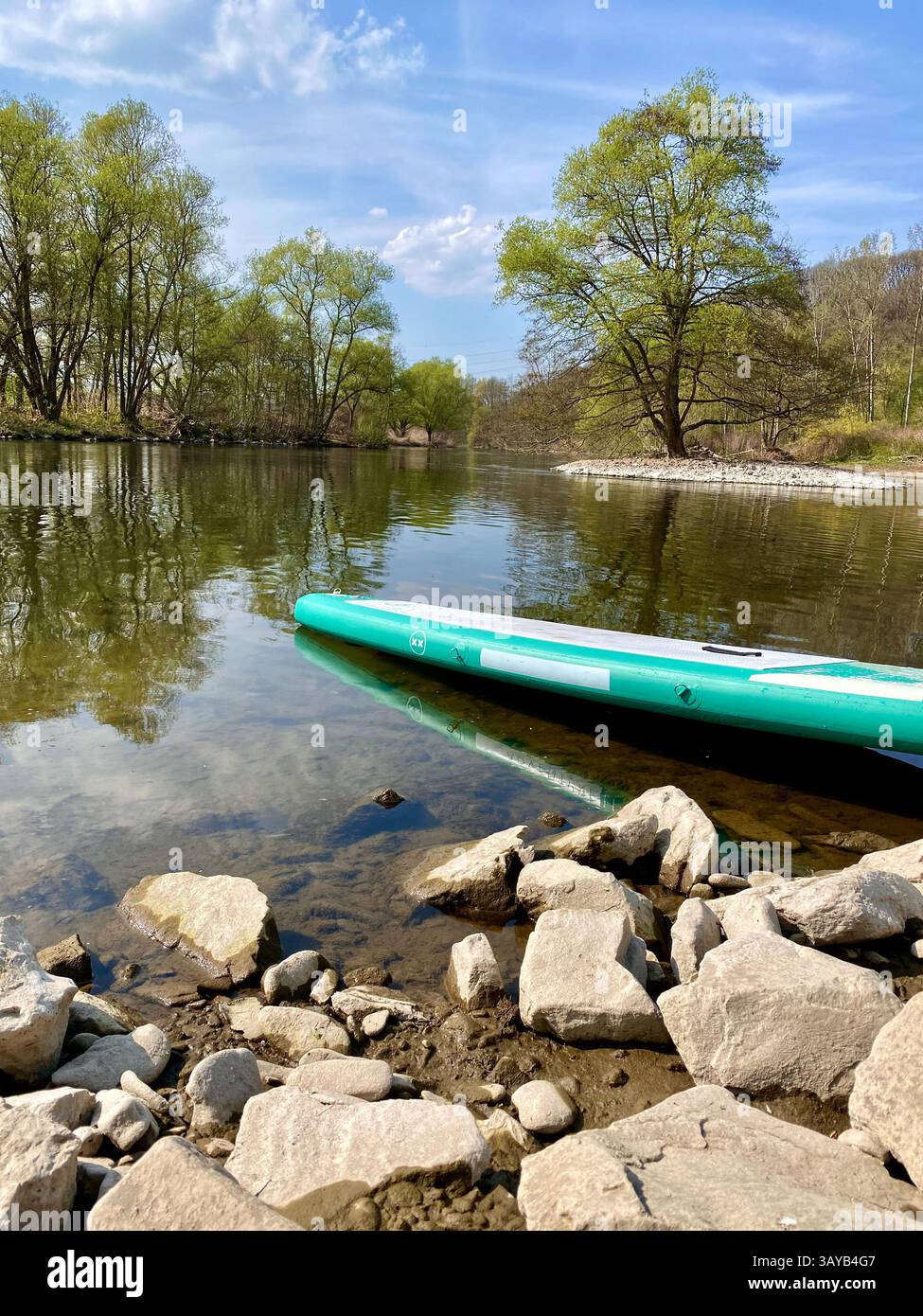 A tranquil spring river scene with crystal-clear water, a rocky riverbank in the foreground, and vibrant green trees lining the opposite shore. A turq - Smartphone Captured Stock Image