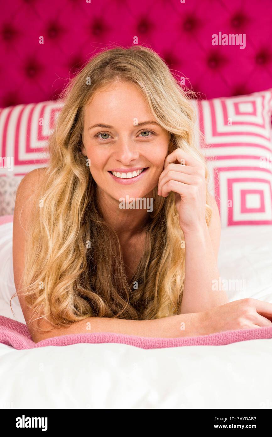 Woman lying on stomach in bedroom, smiling at camera with pink headboard and decorative pillows Stock Photo