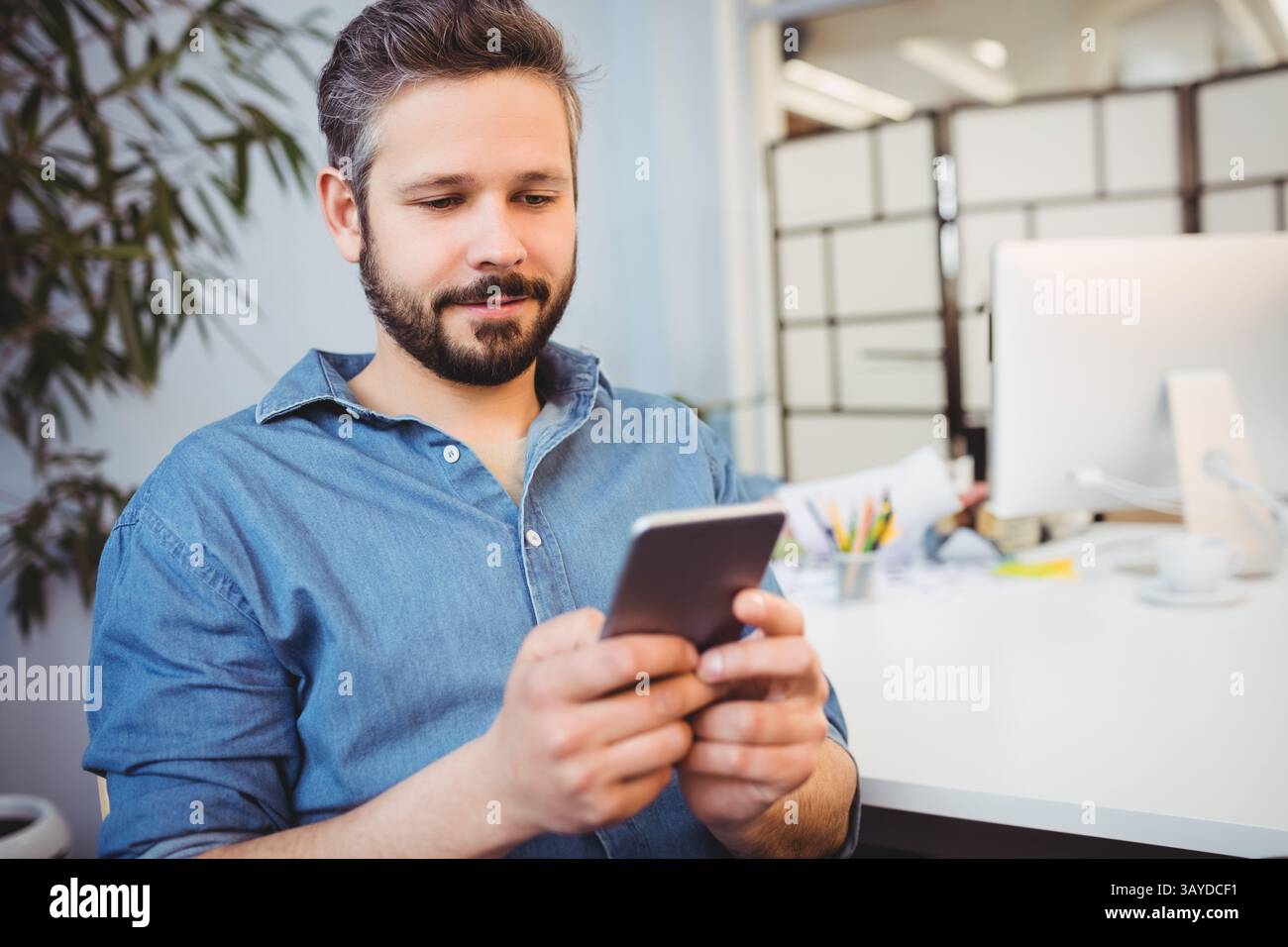 Man scrolling smartphone at modern office desk with monitor, colored pencils, coffee cup and plant Stock Photo