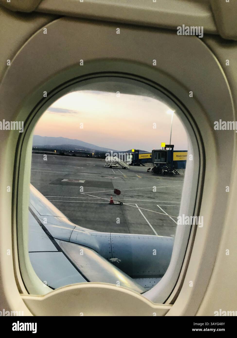 A tranquil sunset view from a Transavia plane at Athens Eleftherios Venizelos Airport, showing the wing, jet bridge with Piraeus Bank logo, and distan - Smartphone Captured Stock Image