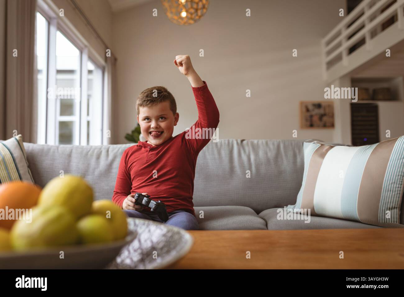 Boy playing video game with black controller while sitting on gray sofa in modern living room Stock Photo