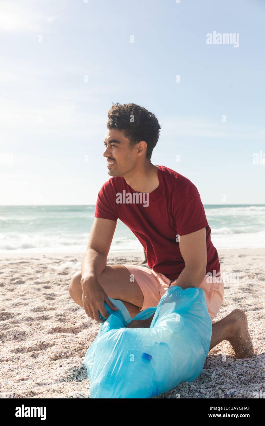Man kneeling on sandy beach near ocean waves, holding blue plastic trash bag while smiling Stock Photo