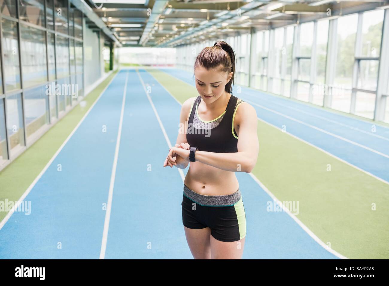 young adult woman checking smartwatch on blue track lanes in gym, wearing sports bra and shorts Stock Photo