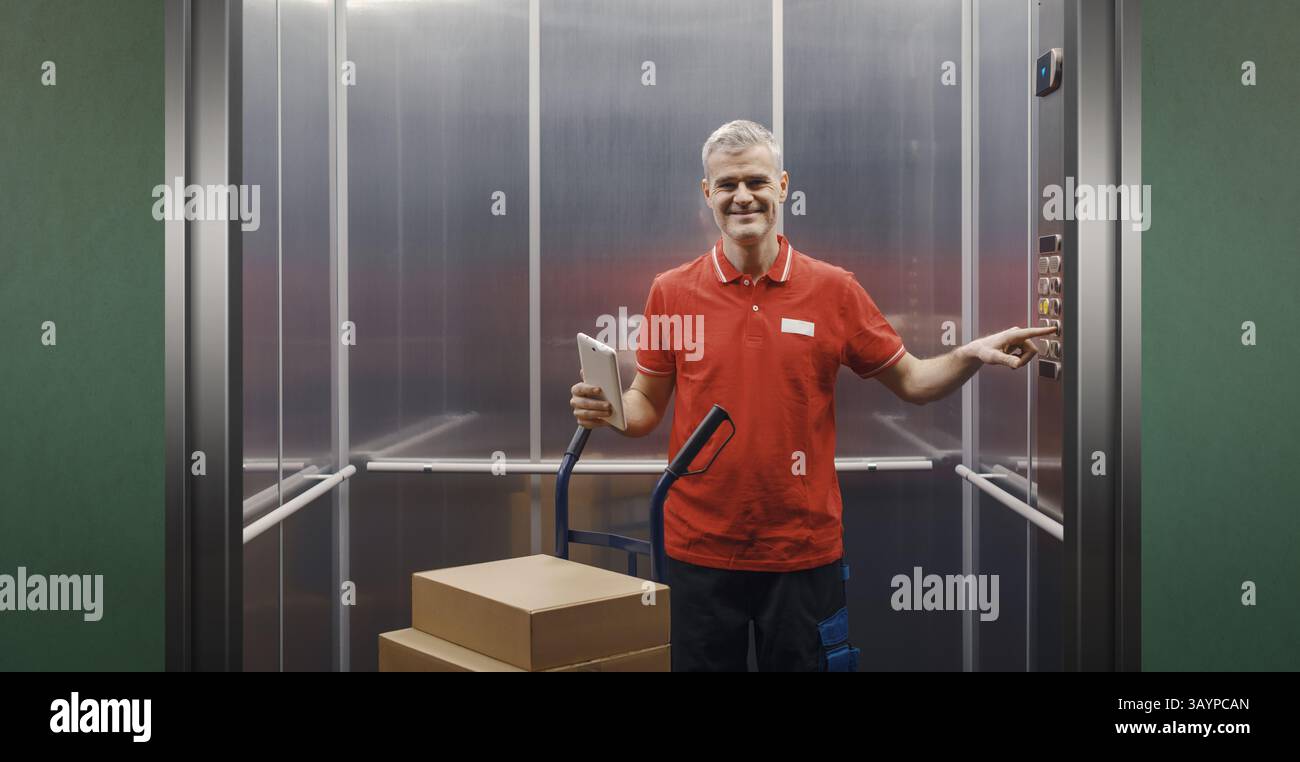 Smiling confident delivery person standing in the lift and pressing a button on the control panel, he is holding a tablet and delivering parcels using Stock Photo