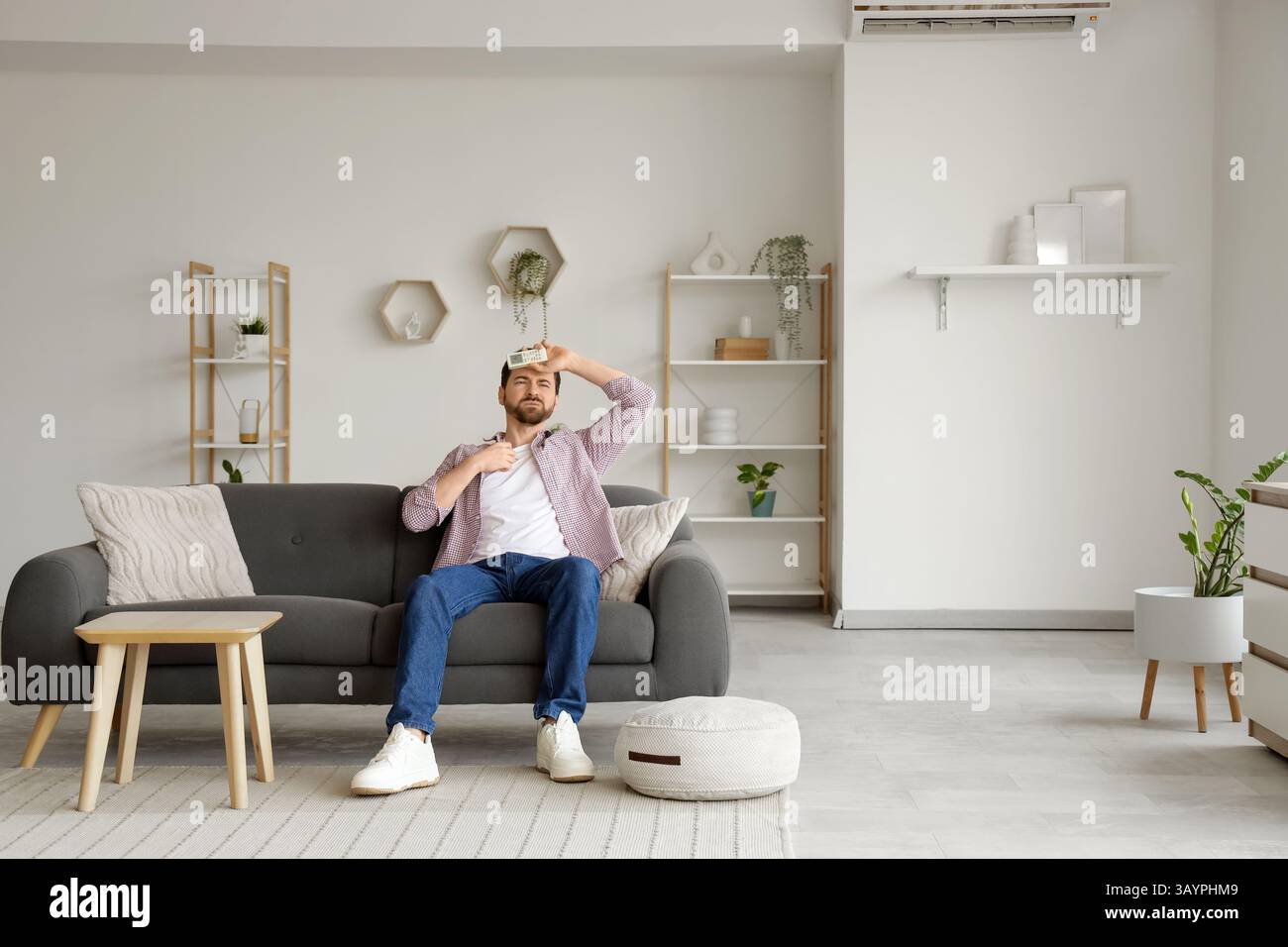 Young man with air conditioner remote control suffering from heat on sofa in living room Stock Photo