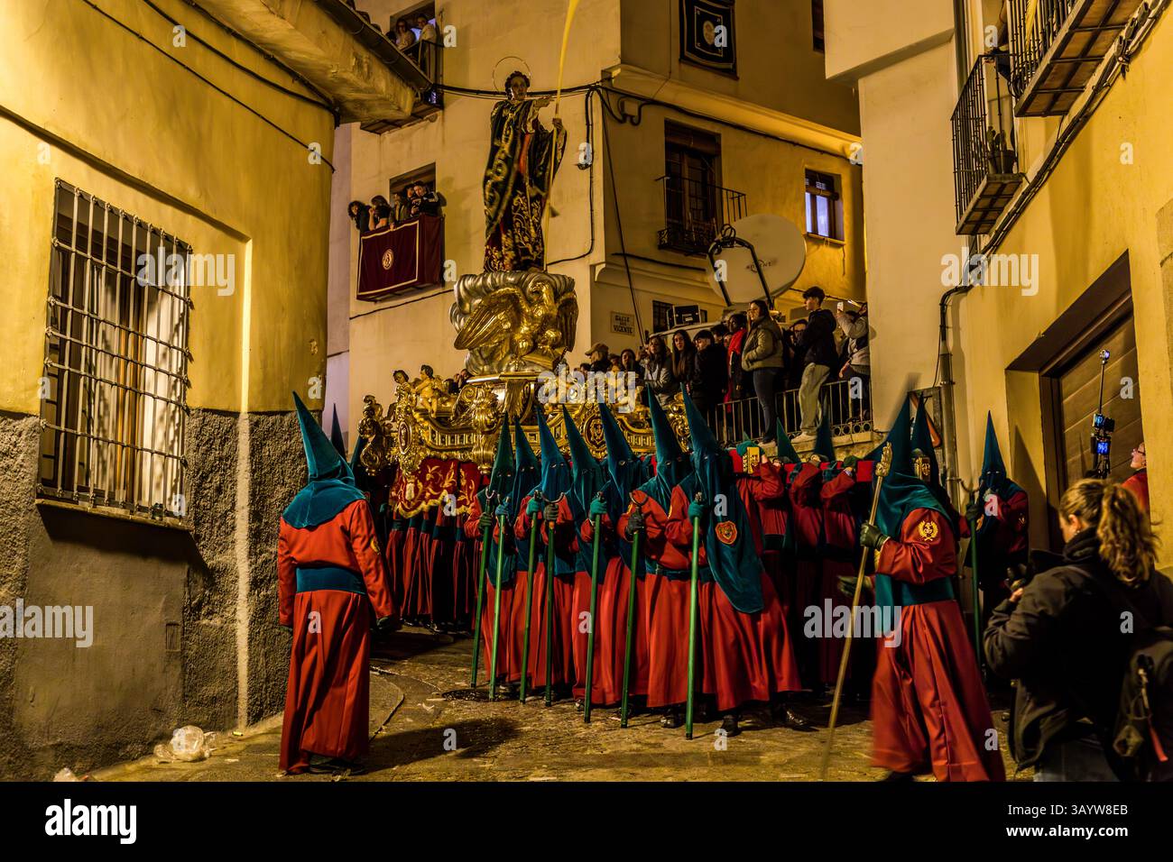 The Venerable Hermandad de San Juan Evangelista has a very special style of movement: its paso depicts the favourite disciple of Jesus Christ and is not simply carried forward but given a special dynamic through rhythmic rocking or swaying ("bamboleo") and a slightly springy motion. This movement is meant to symbolise the youth and hope associated with the apostle John. Calle Alonso de Ojeda, Cuenca, Castilla-La Mancha, Spain. Bearers in red and blue robes carry the paso while spectators watch from balconies. During Semana Santa in Cuenca, 33 Hermandades take part in eleven processions through Stock Photo