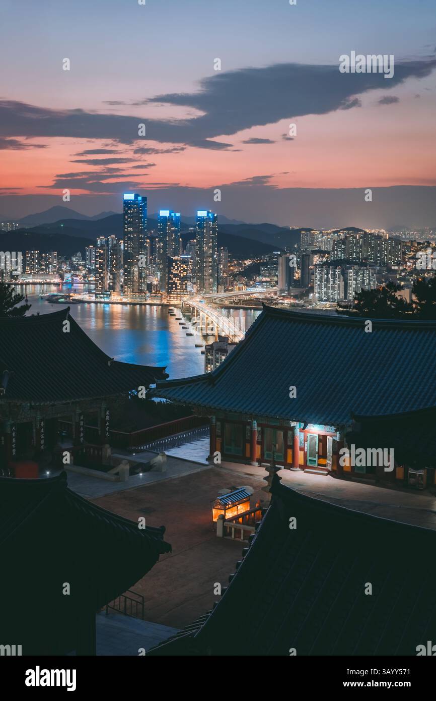 Evening cityscape of Busan, South Korea with traditional temple in foreground and skyscrapers illuminated along the coastline. Stock Photo