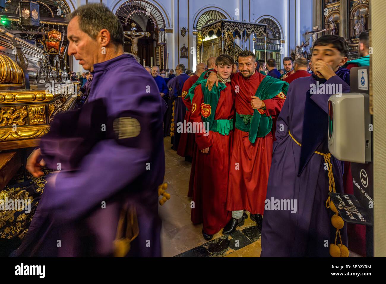In the parish church of El Salvador, members of various brotherhoods prepare for the long procession, which begins at 5.30 am with a kind of gauntlet run. Plaza Salvador, Cuenca, Castilla-La Mancha, Spain. Bearers in purple robes stand beside two young men in red robes with green sashes. During Semana Santa in Cuenca, 33 Hermandades take part in eleven processions through the city during Holy Week. Stock Photo