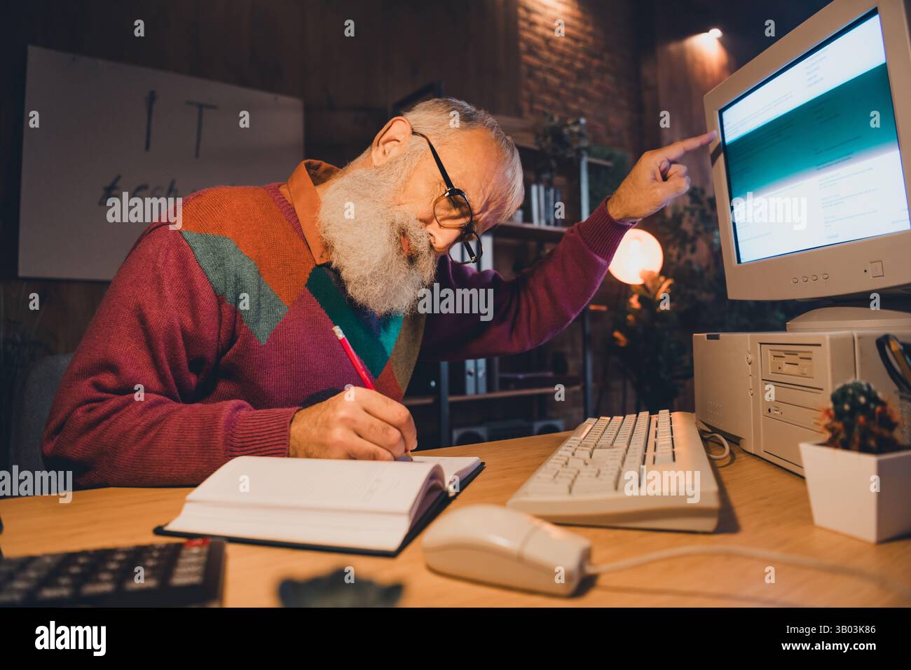 Elderly programmer working attentively at his desk in cozy evening light, taking notes and analyzing data on desktop computer Stock Photo