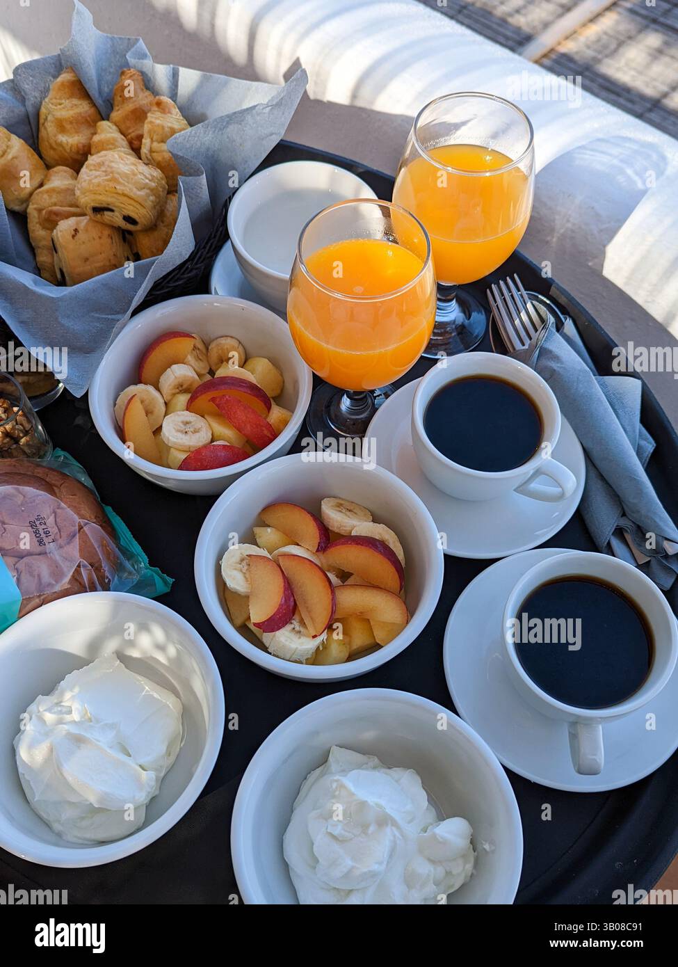 A continental breakfast featuring coffee, orange juice, fresh fruit and yoghurt, meats and cheese, and pastries on a tray outside on a hotel balcony. - Smartphone Captured Stock Image