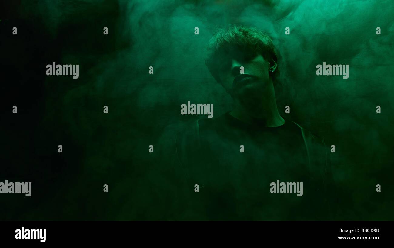 Young man in dark shirt and leather coat stands surrounded by green mist in moody atmosphere against dark studio background Stock Photo