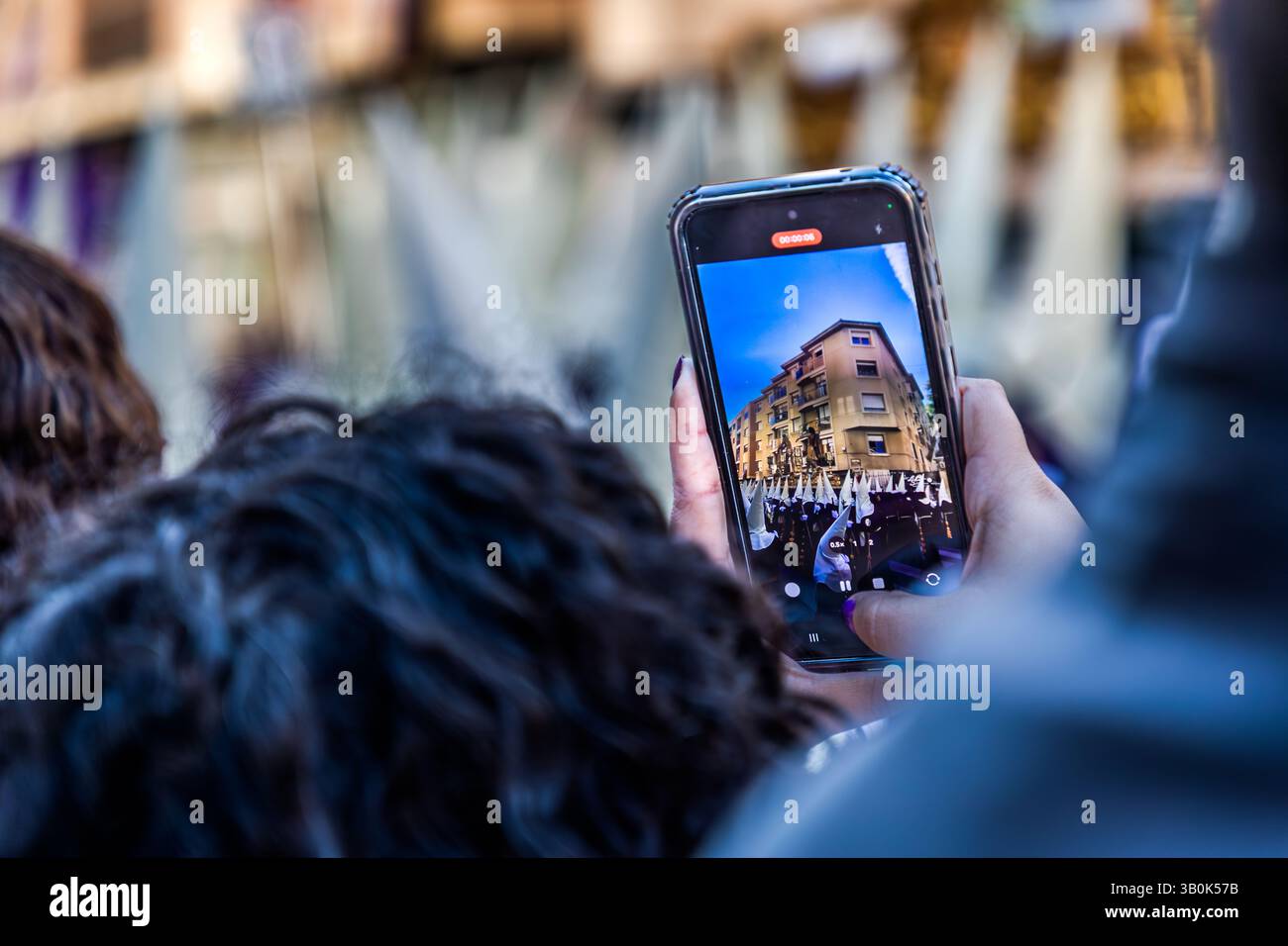 A bystander records a Maundy Thursday procession with a smartphone as the Hermandad de Nuestro Padre Jesús Amarrado a la Columna passes in Parque del Huécar, Cuenca, Castilla-La Mancha, Spain. The phone's screen shows the procession moving past the town's ochre-coloured houses. Stock Photo