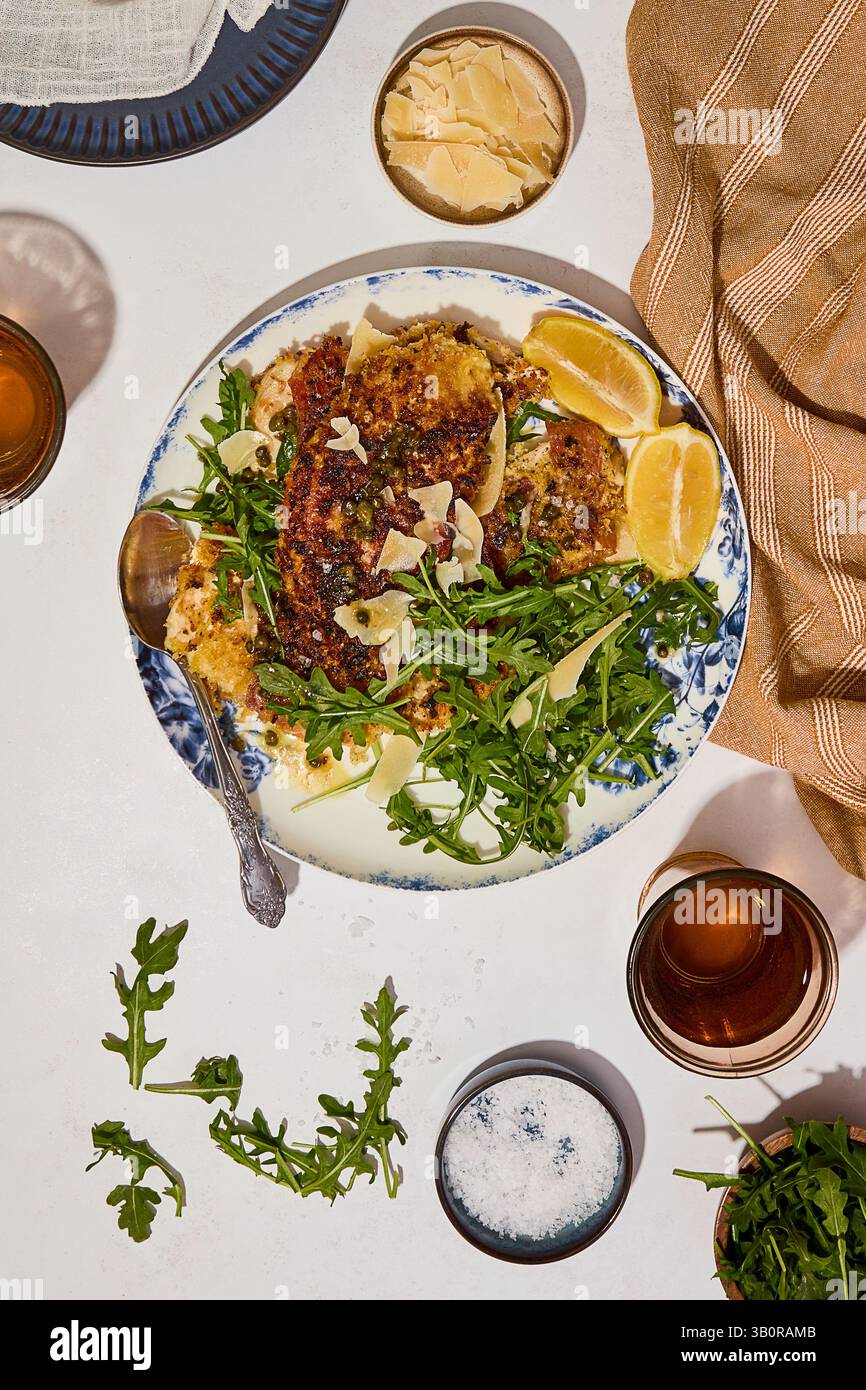 A blue-rimmed plate with crispy chicken cutlet topped with arugula, parmesan, and lemon wedges, surrounded by drinks and condiments. Stock Photo