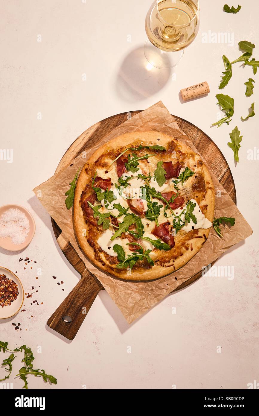 Artisanal prosciutto and arugula pizza on wooden board with white wine, fresh herbs, and seasoning dishes in elegant setting. Stock Photo