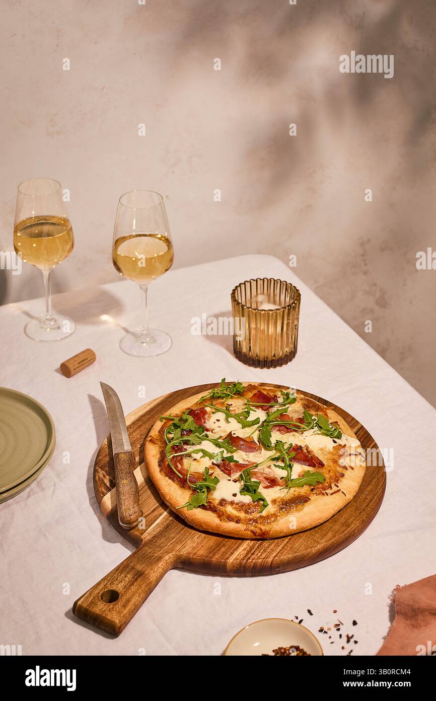 Elegant dining setup with prosciutto-arugula pizza on wooden board, white wine glasses, candle holder, and knife on textured background. Stock Photo
