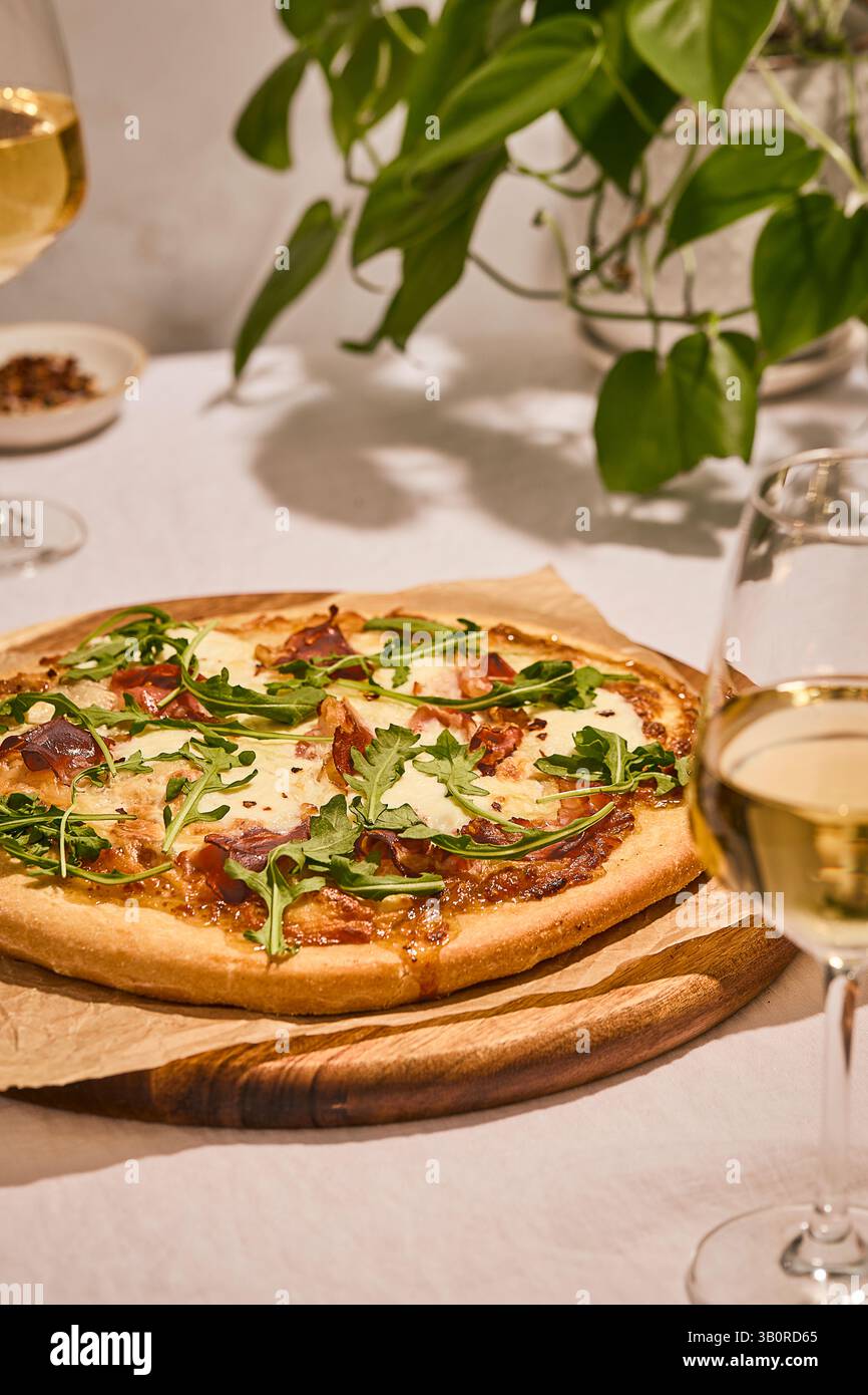 Rustic prosciutto-arugula pizza on wooden board with white wine glasses and houseplant, creating an elegant dining atmosphere. Stock Photo