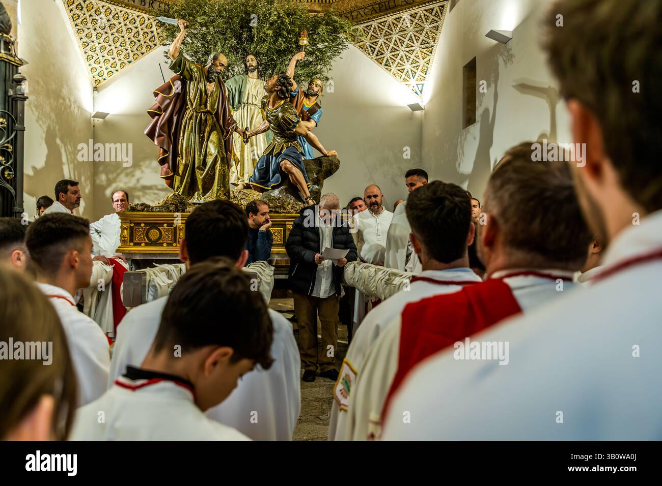 Members of the Hermandad de San Pedro Apóstol gather for the roll call in the Iglesia de San Pedro in Cuenca, Castilla-La Mancha, Spain, before the Procesión del Silencio begins. An elderly man reads out a list while the members, dressed in white and red, stand around him. In the background, the paso showing Peter and the servant Malchus is visible. Calle Trabuco, Cuenca, Castilla-La Mancha, Spain. Stock Photo