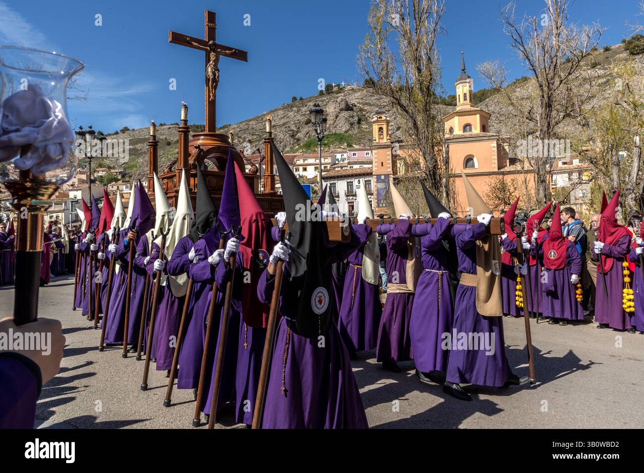 Penitents of the Archicofradía de Paz y Caridad, wearing purple robes and hoods, carry a large wooden crucifix through Calle San Lázaro in Cuenca, Castilla-La Mancha, Spain. The independent and very old brotherhood unites several hermandades and stages its own procession on Maundy Thursday, in which members of all affiliated hermandades take part in their respective habit colours. It is one of 33 hermandades that stage a total of eleven processions during Cuenca's Semana Santa. Stock Photo