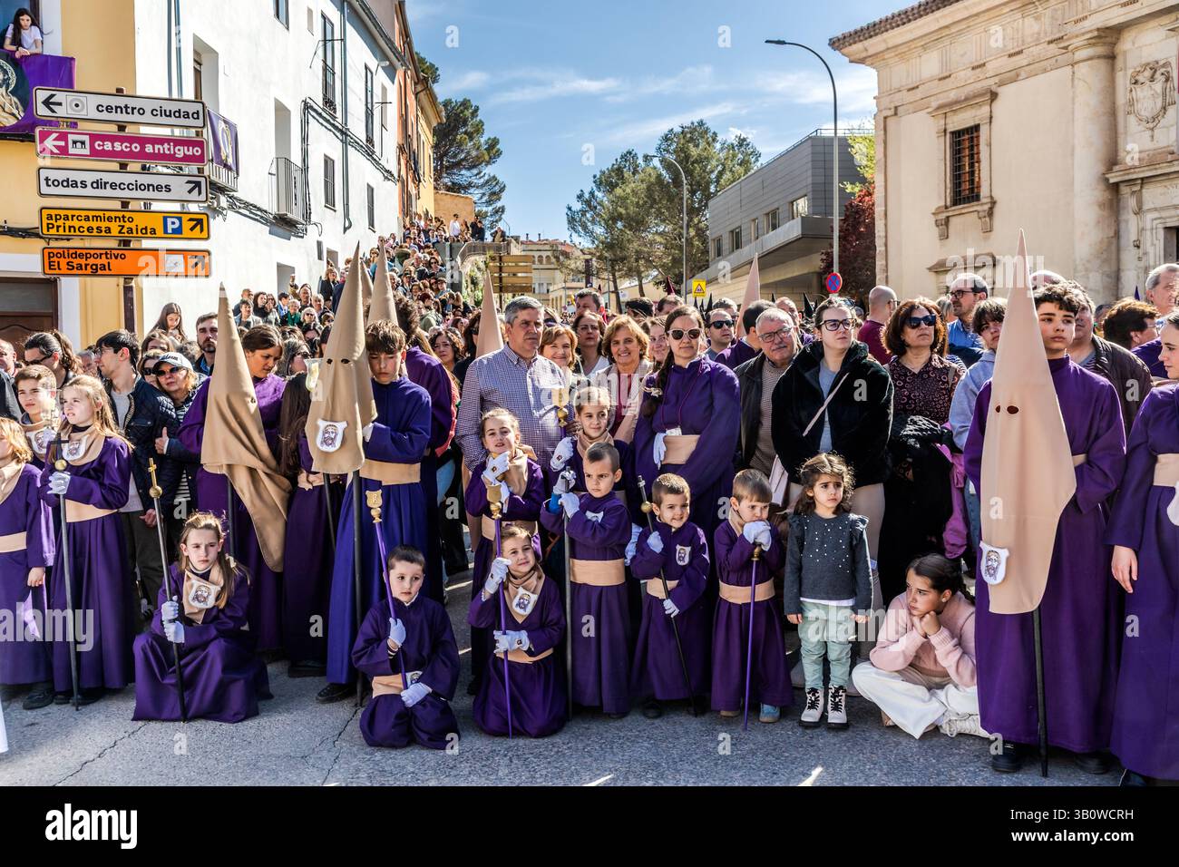 Large crowds follow the Maundy Thursday procession on Calle San Lázaro in Cuenca, Castilla-La Mancha, Spain. In the foreground, children in purple robes and cream-coloured hoods kneel as participants in the procession. Cuenca's Semana Santa involves 33 hermandades that stage a total of eleven processions. Stock Photo