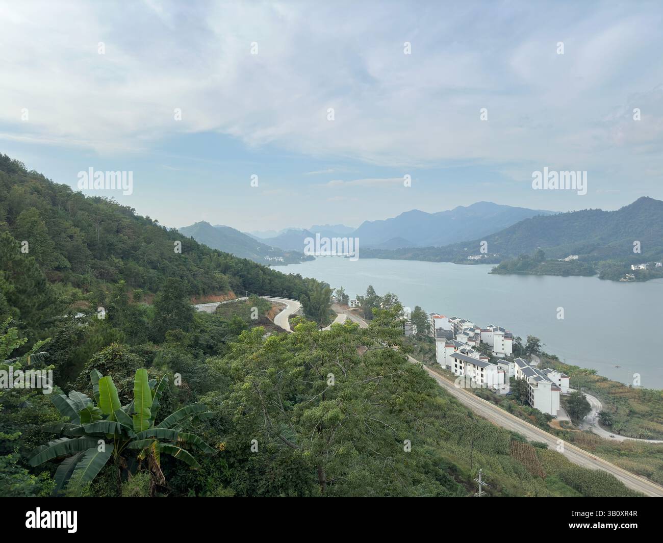 A winding road leads towards houses near a serene lake with beautiful mountain views. - Smartphone Captured Stock Image