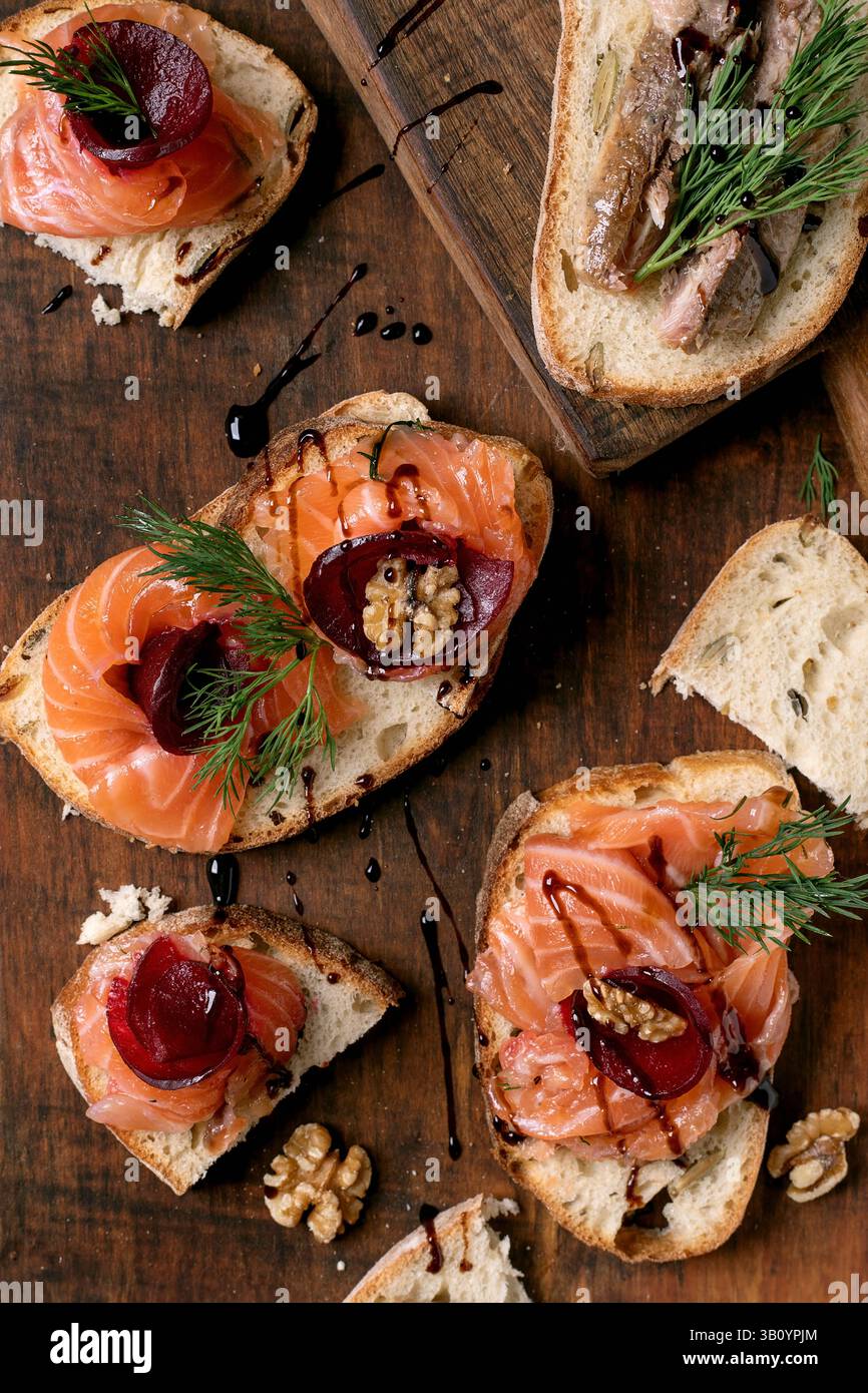 Sandwiches with thinly sliced salted salmon fillet, sardines, beetroot slices, walnuts, balsamic vinegar and fresh wheat bread on wooden board. Brown Stock Photo