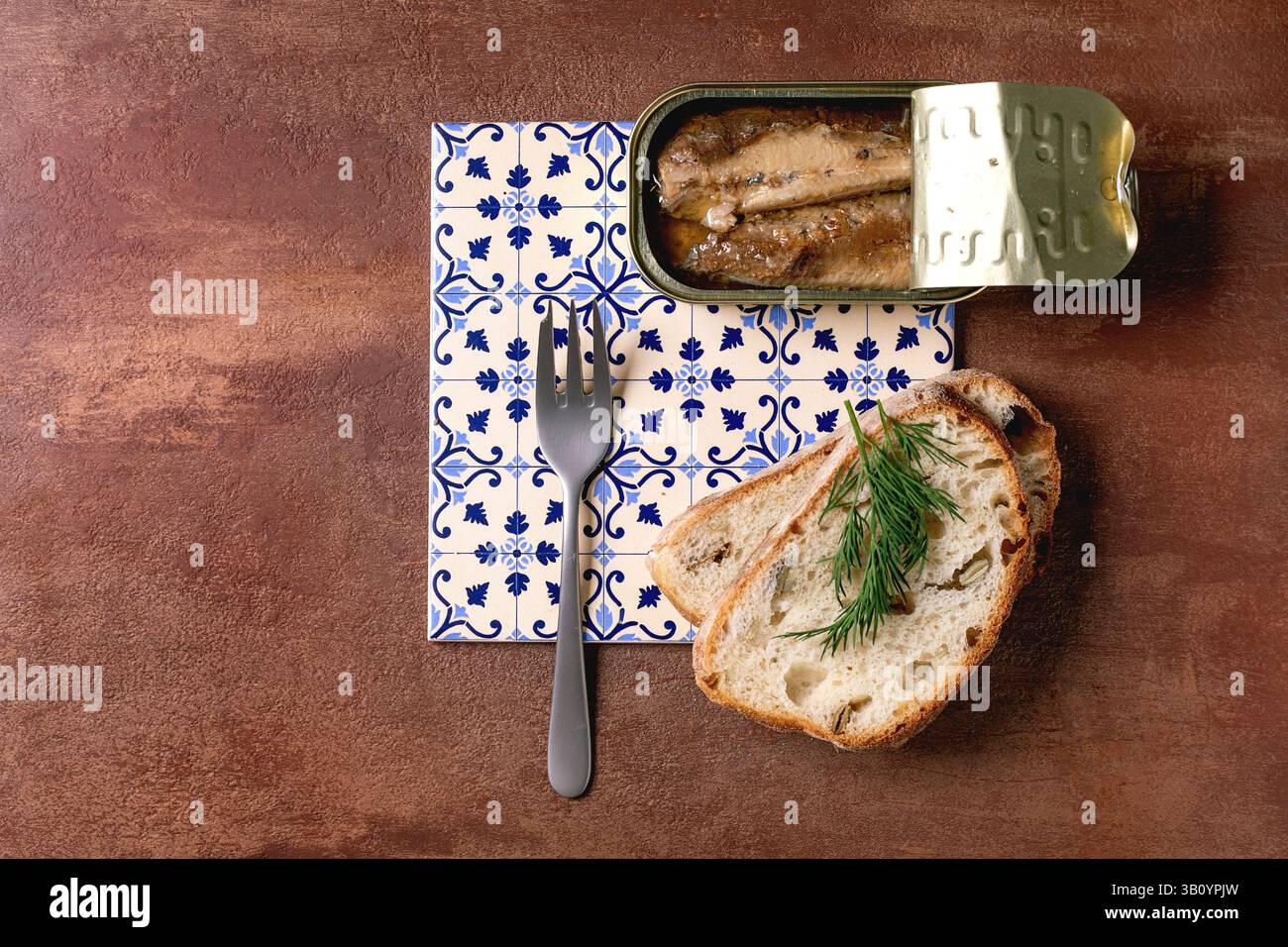 Open tin of sardine fillets in oil, served with fresh wheat bread, dill, and fork on white-and-blue patterned ceramic tiles. Textured brown background Stock Photo