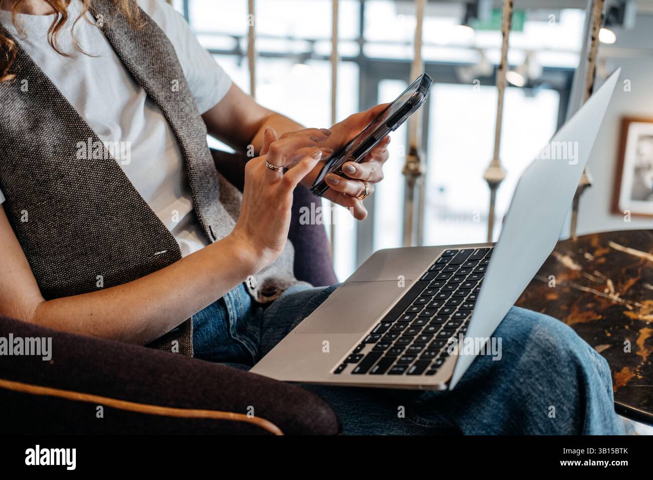 A close-up of a young womans hands as she interacts with her smartphone while working on a laptop. Cross-platform work habits, device integration Stock Photo