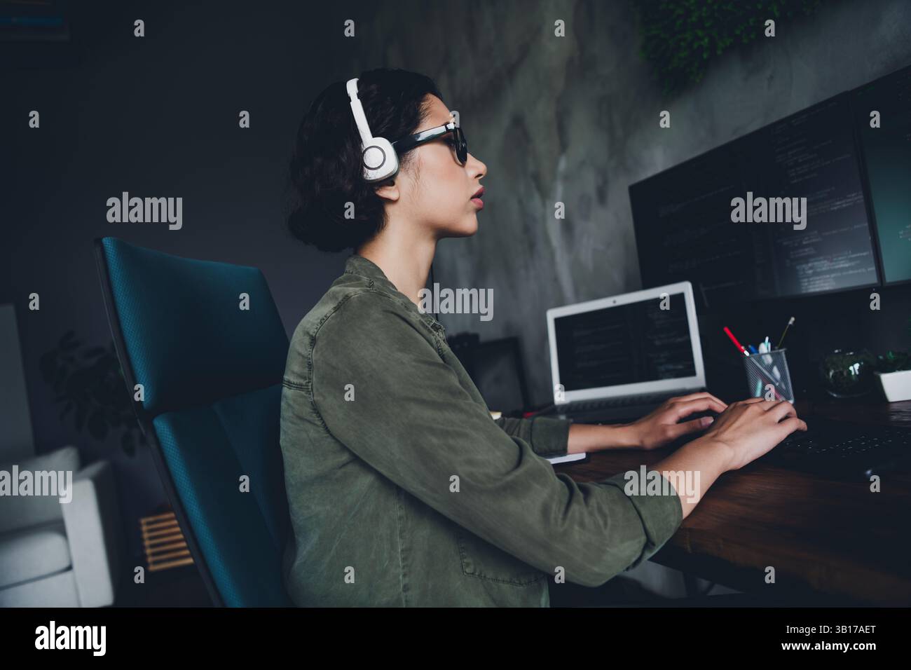 Young female coder working in a modern office environment with programming code on computers, wearing headphones, focused on programming Stock Photo