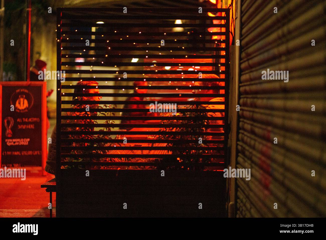 Two people sit in a warmly lit outdoor seating area of a bar or restaurant at night, partially obscured by a slatted screen and plants. Stock Photo