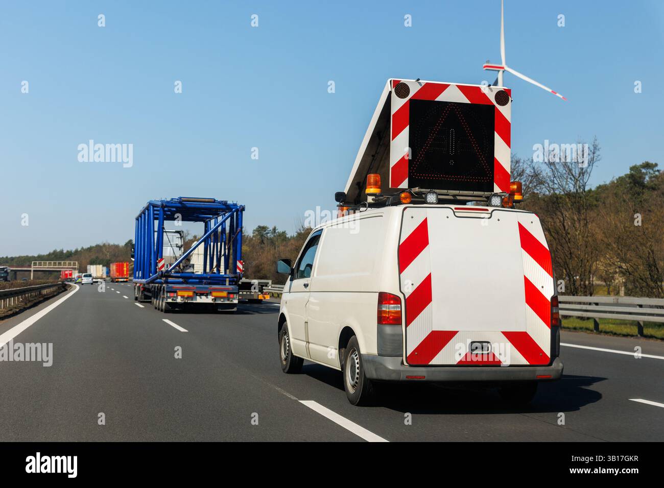 Escort van with warning sign follows a heavy-duty truck transporting large blue construction frame on multilane highway in Germany. White special Stock Photo