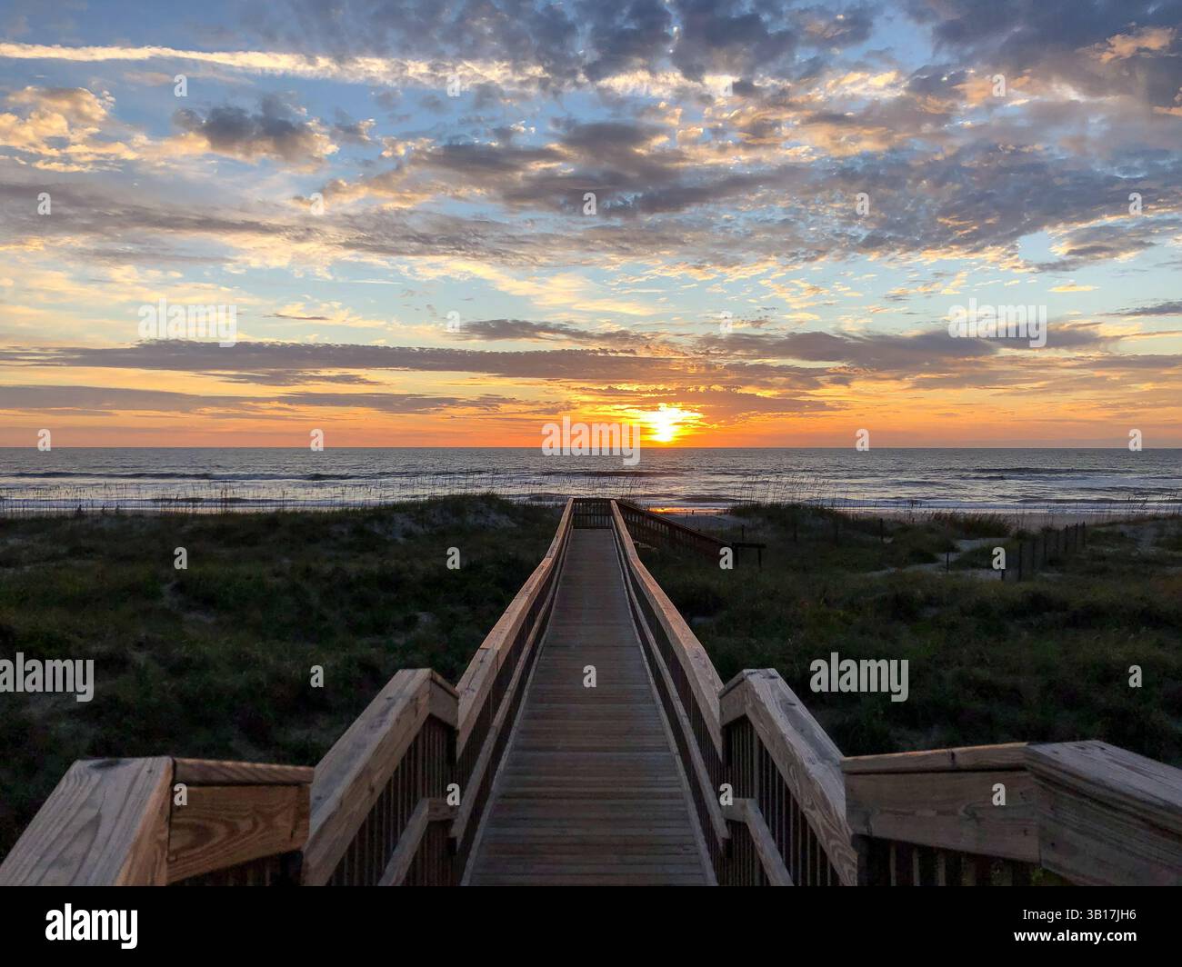 A view of a wooden boardwalk leading to the beach with sun peaking through the clouds, colorful sky and ocean in the background. Amelia Island, Florid - Smartphone Captured Stock Image