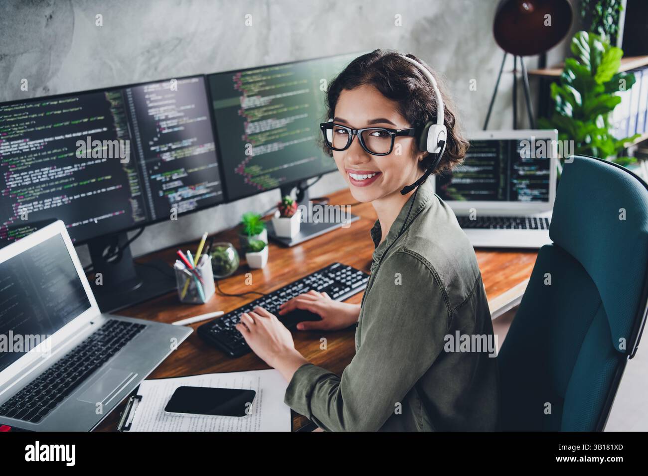 Young female software developer working remotely on a programming project, smiling confidently in a modern home office environment Stock Photo