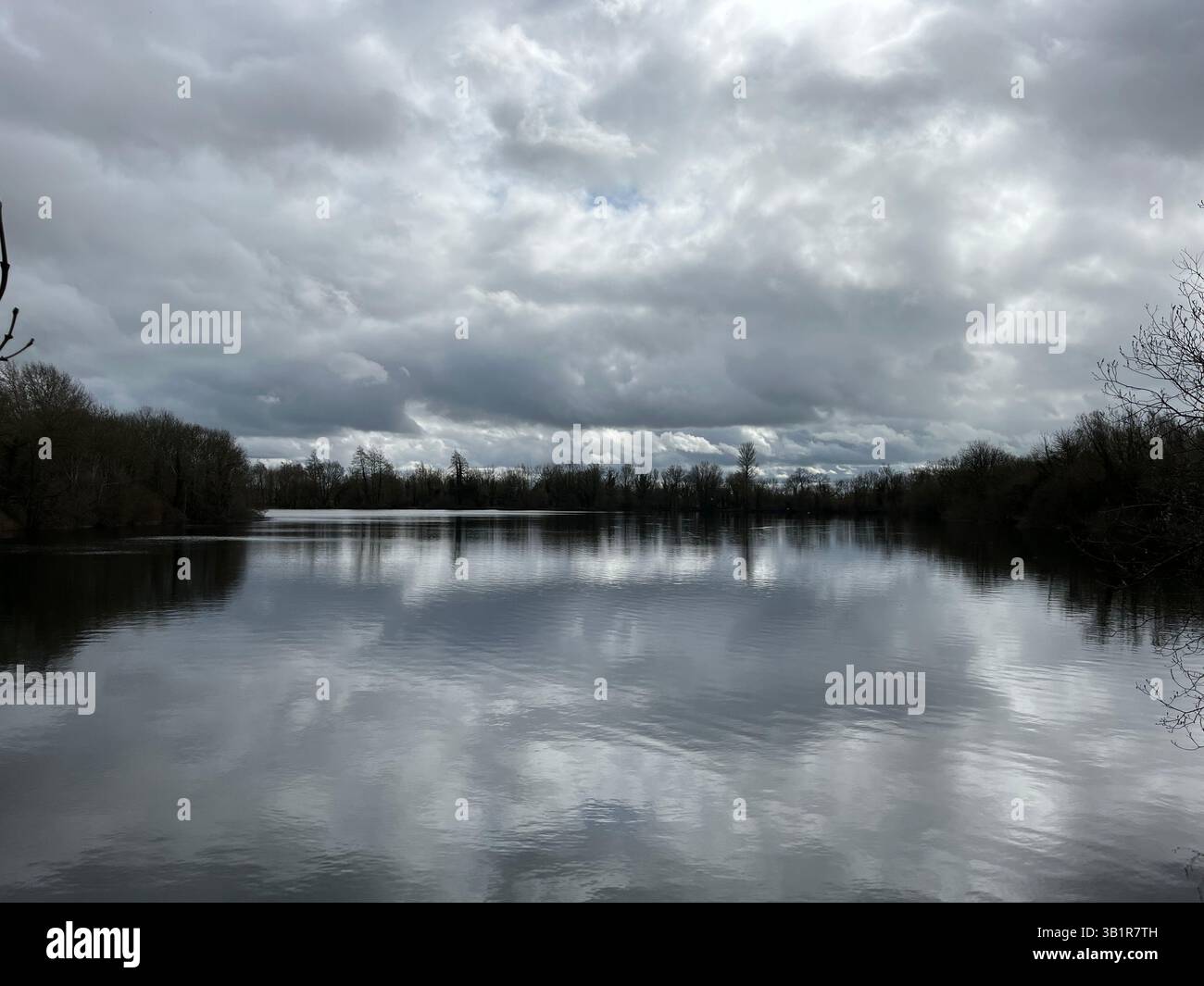 Image of lake on on the Cotswold Water Park on a grey and cloudy day, reflection of clouds and tree line. Eerie, isolation, beauty. - Smartphone Captured Stock Image
