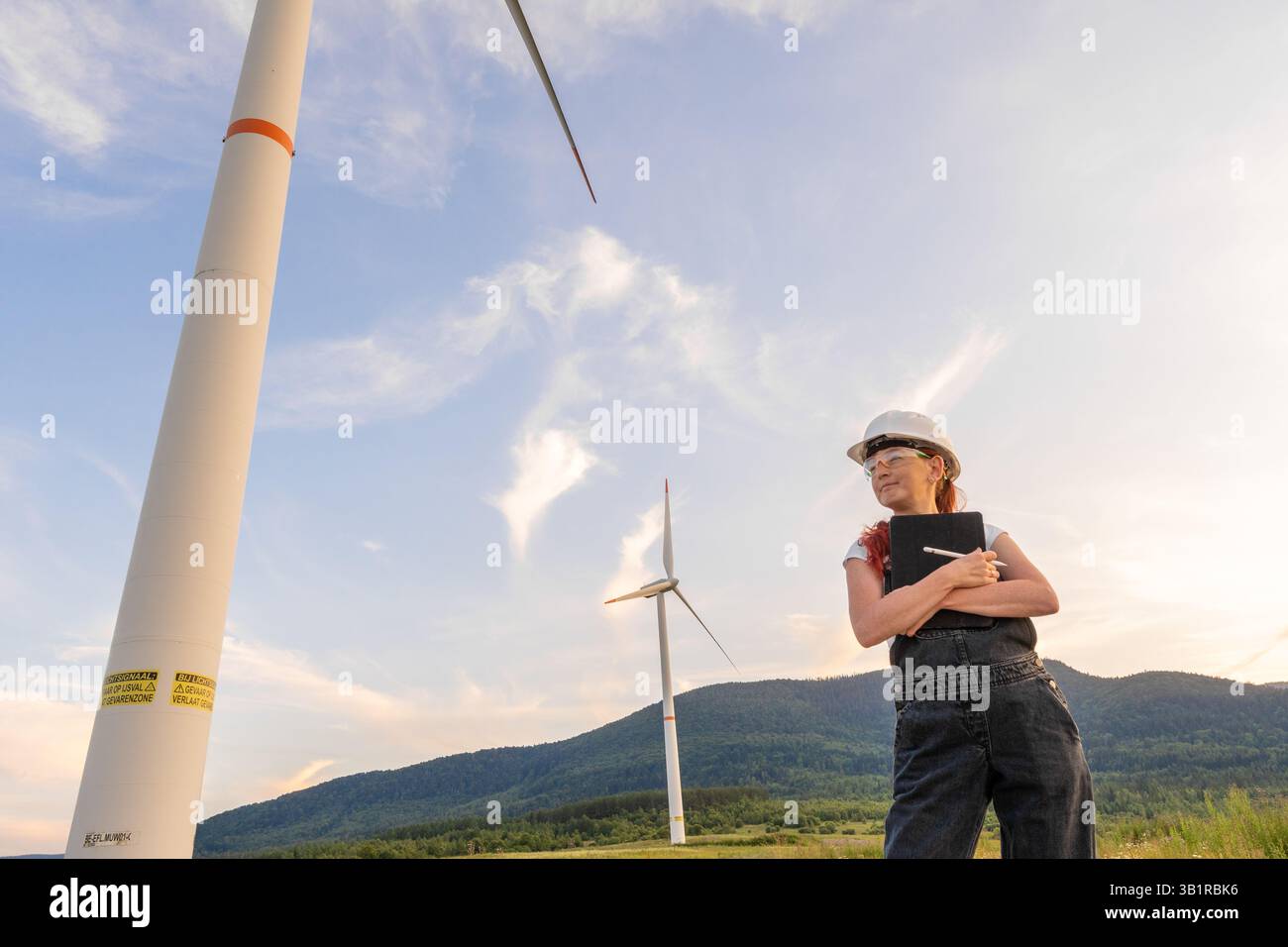 A confident female engineer stands beside a tall wind turbine ...