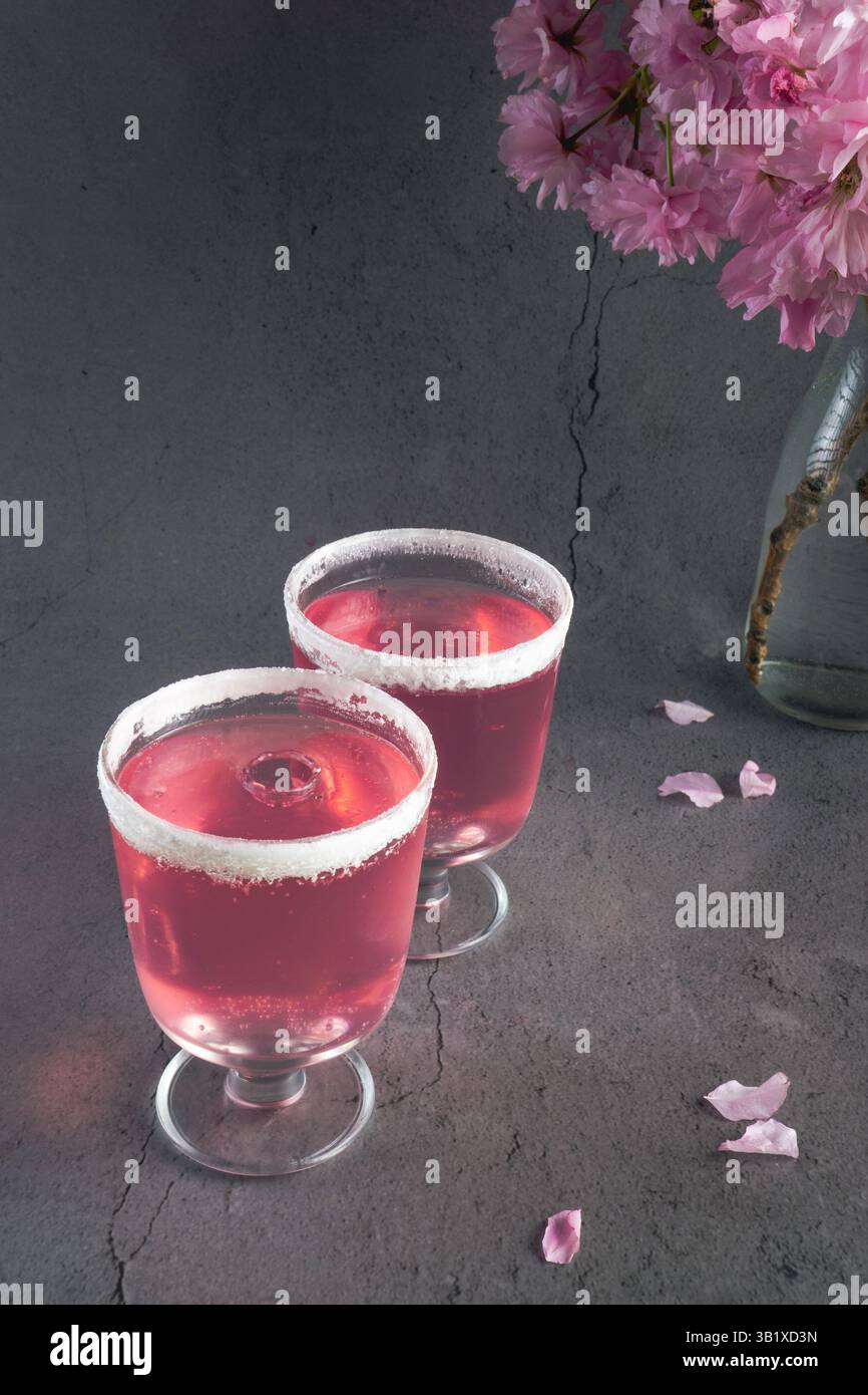 two small glasses filled with a pink drink, rimmed with sugar, sitting on a dark surface with fallen pink cherry blossom petals Stock Photo